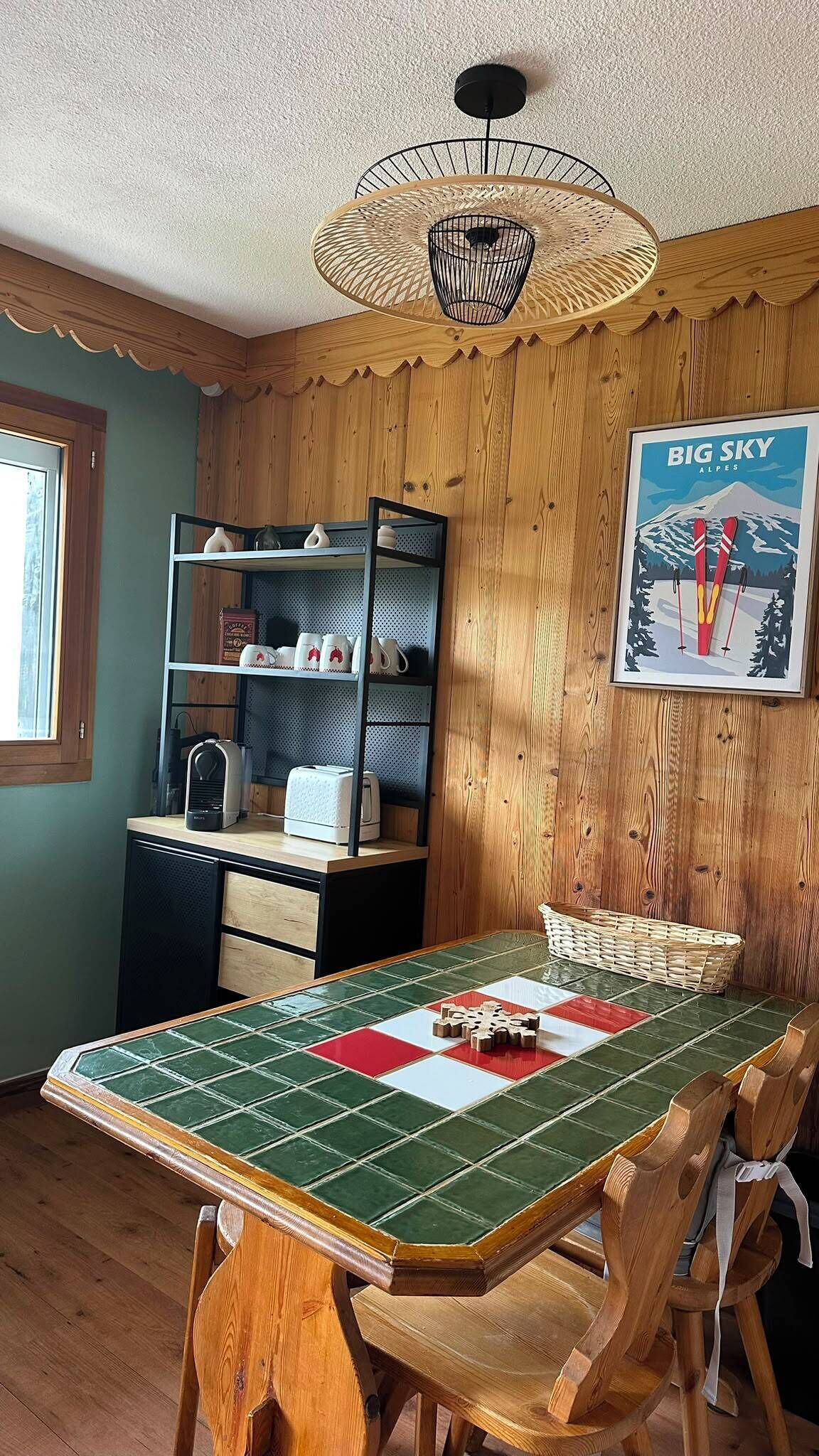 Un comedor con mesa de madera, sillas, una lámpara de techo tejida, una zona de cocina con estantes y un cuadro en la pared.