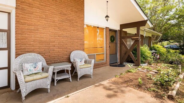 Un edificio de ladrillo con un porche cubierto, dos sillas de mimbre, una mesa pequeña y un jardín con plantas y una valla de madera.