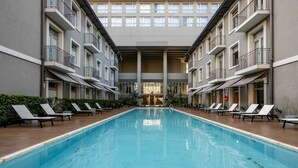 Zona de piscina amb gandules, una piscina central i un edifici de diversos pisos amb balcons.