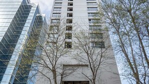 Un edifici alt d'un disseny modern, amb una entrada destacada i portes de vidre, i una zona d'aparcament al davant.