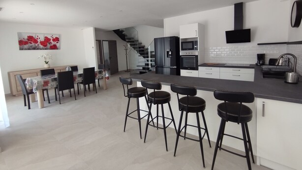 Uma cozinha moderna com uma ilha central, banquetas de bar, área de refeições com cadeiras e uma escada que leva a outro andar.