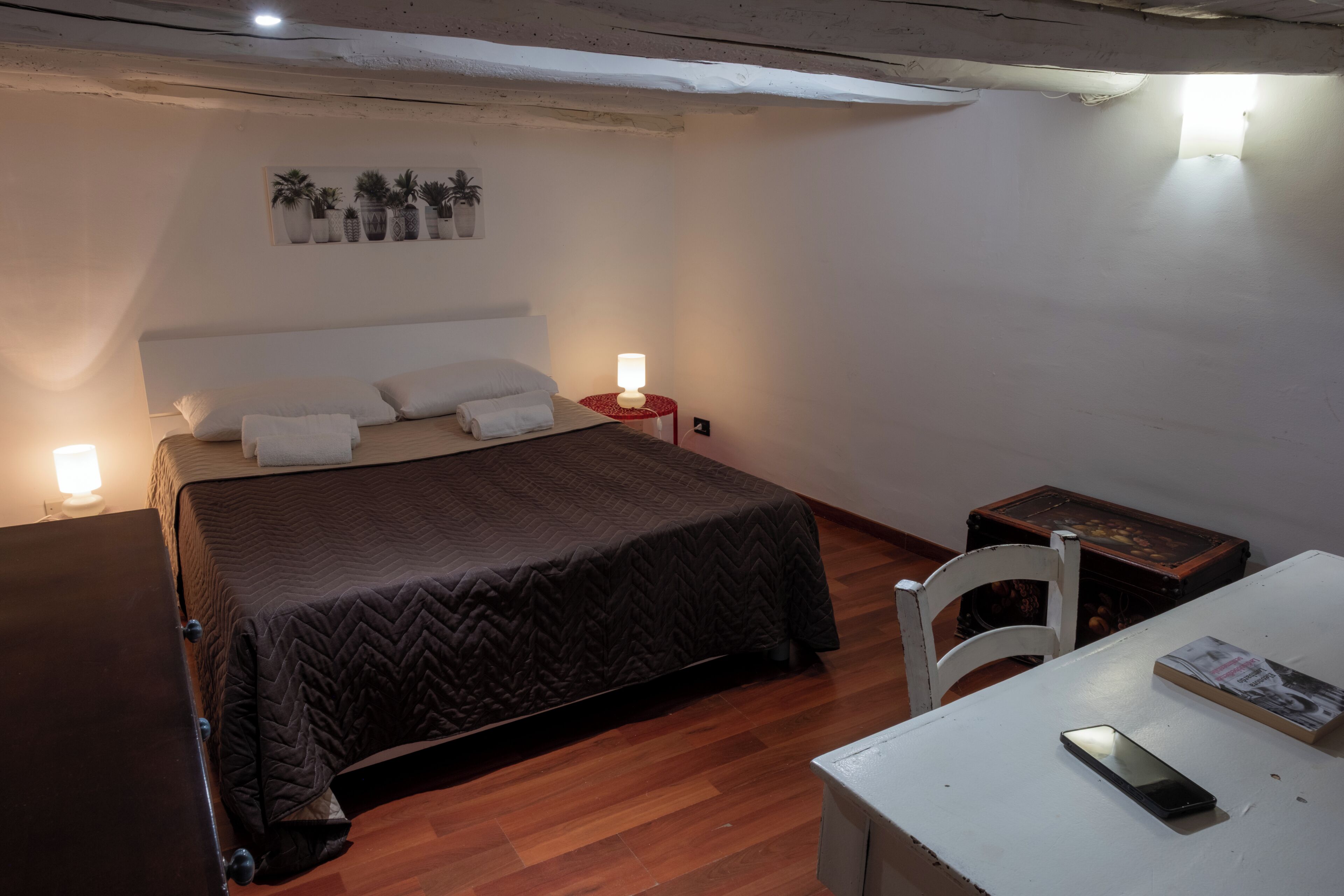 A bedroom with a bed, bedside lamps, a wooden floor, a small table with a book and a phone, and a wall-mounted picture of plants.