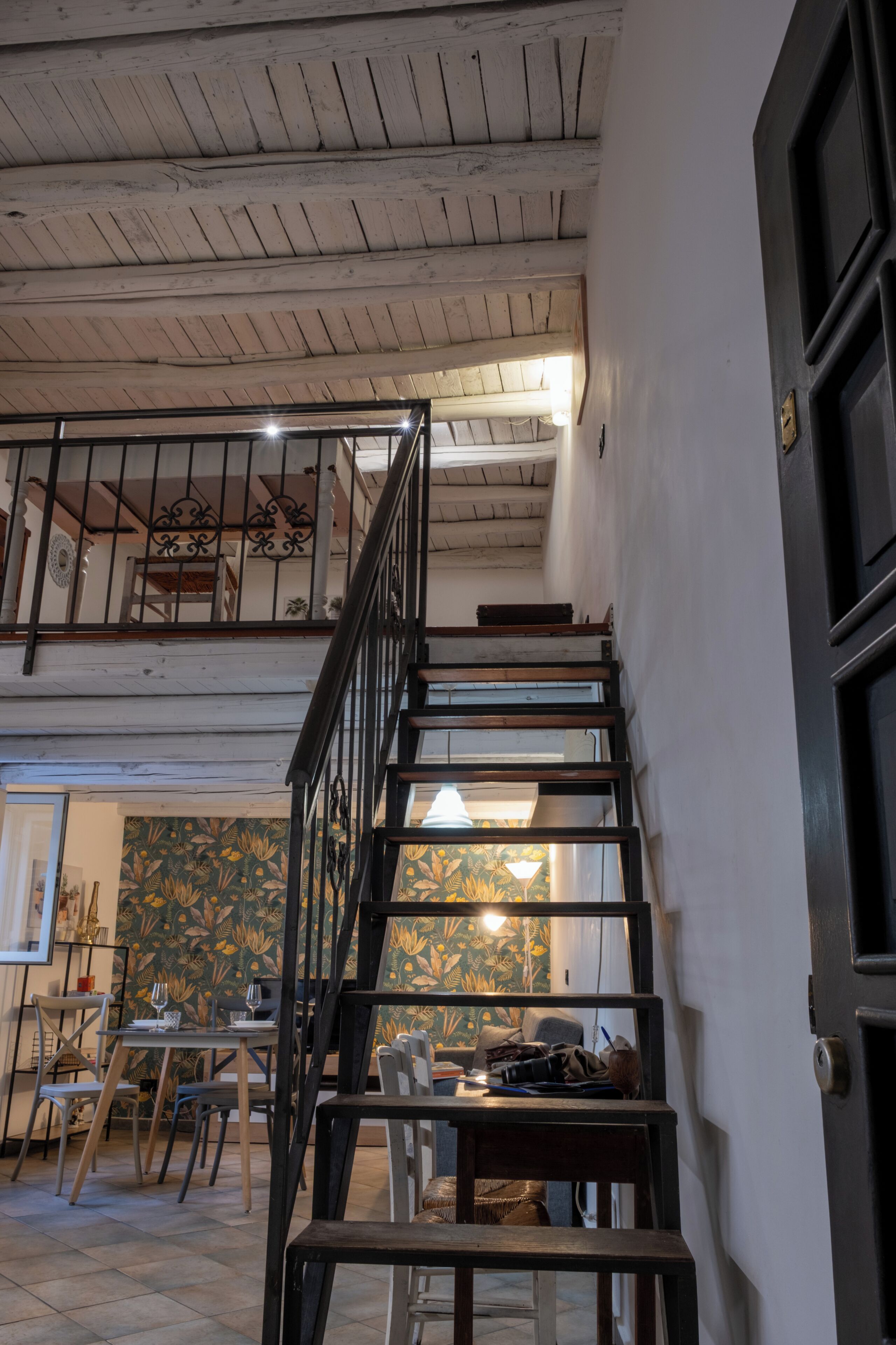 A staircase leading to a second floor with a railing, a dining area with tables and chairs, and a patterned wall.