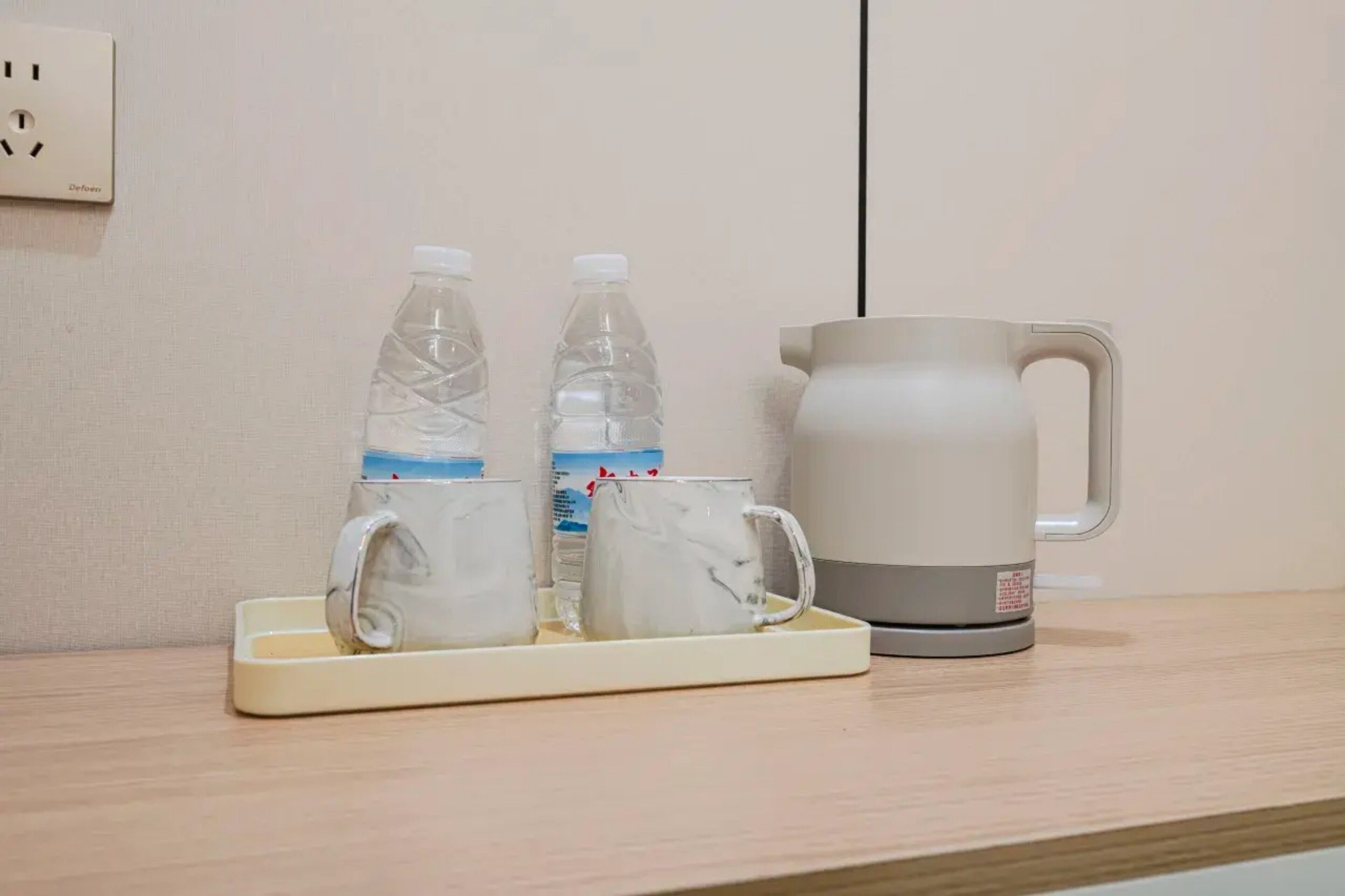 Una bandeja con dos botellas de agua y dos vasos sobre una superficie de madera, al lado de una tetera eléctrica blanca.