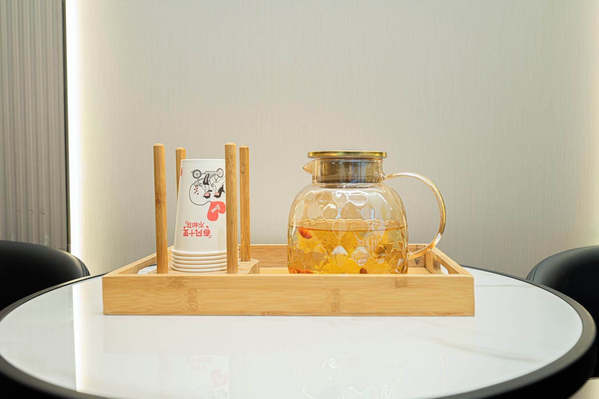 A table with a glass decanter and a stack of white cups with a logo, placed on a wooden tray.