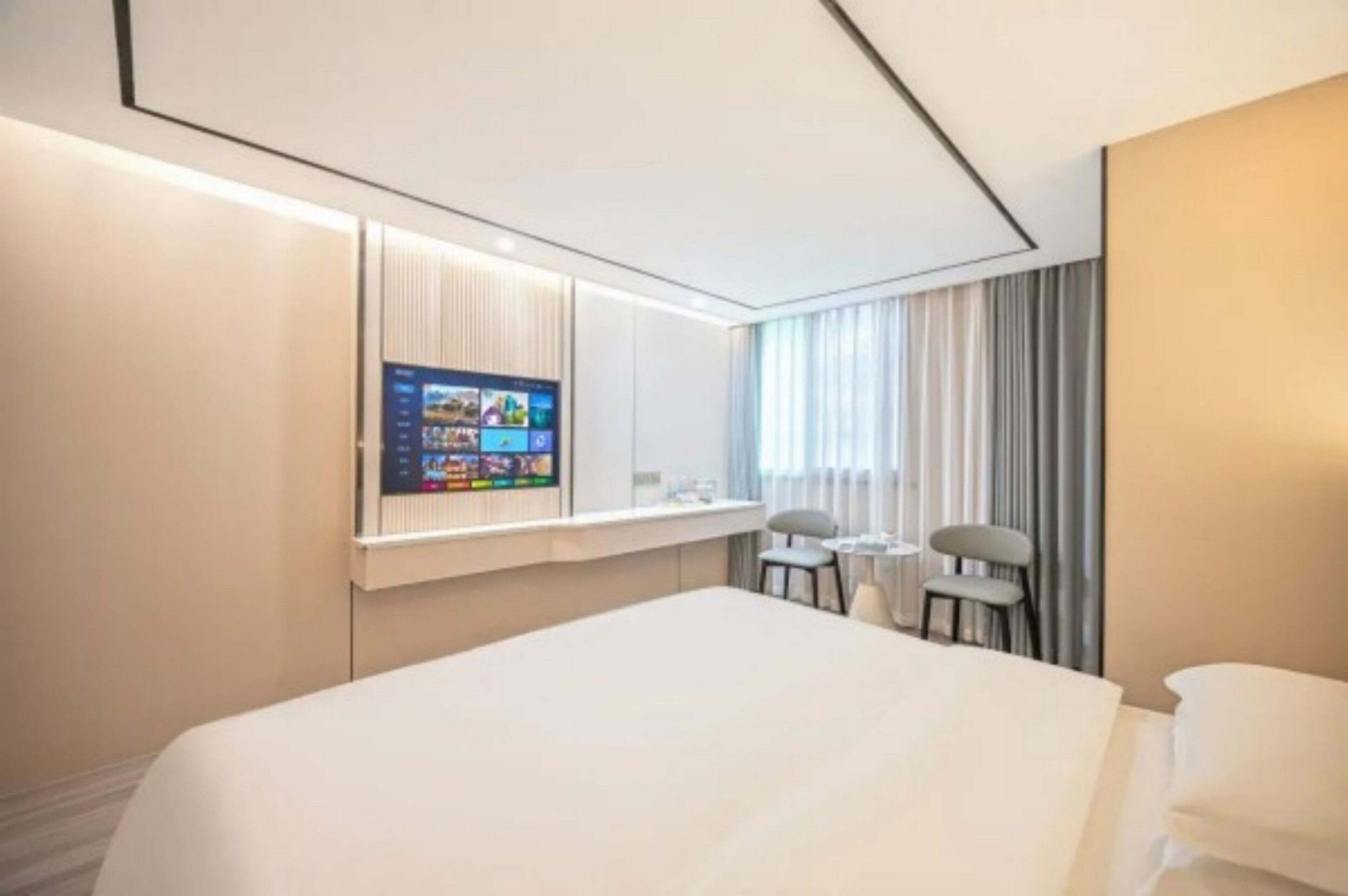 A hotel room with a large bed, a desk with two chairs, a flat-screen TV mounted on the wall, and a window with curtains.