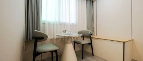 A small, modern room with a round table, two chairs, a desk, and a window with sheer curtains.