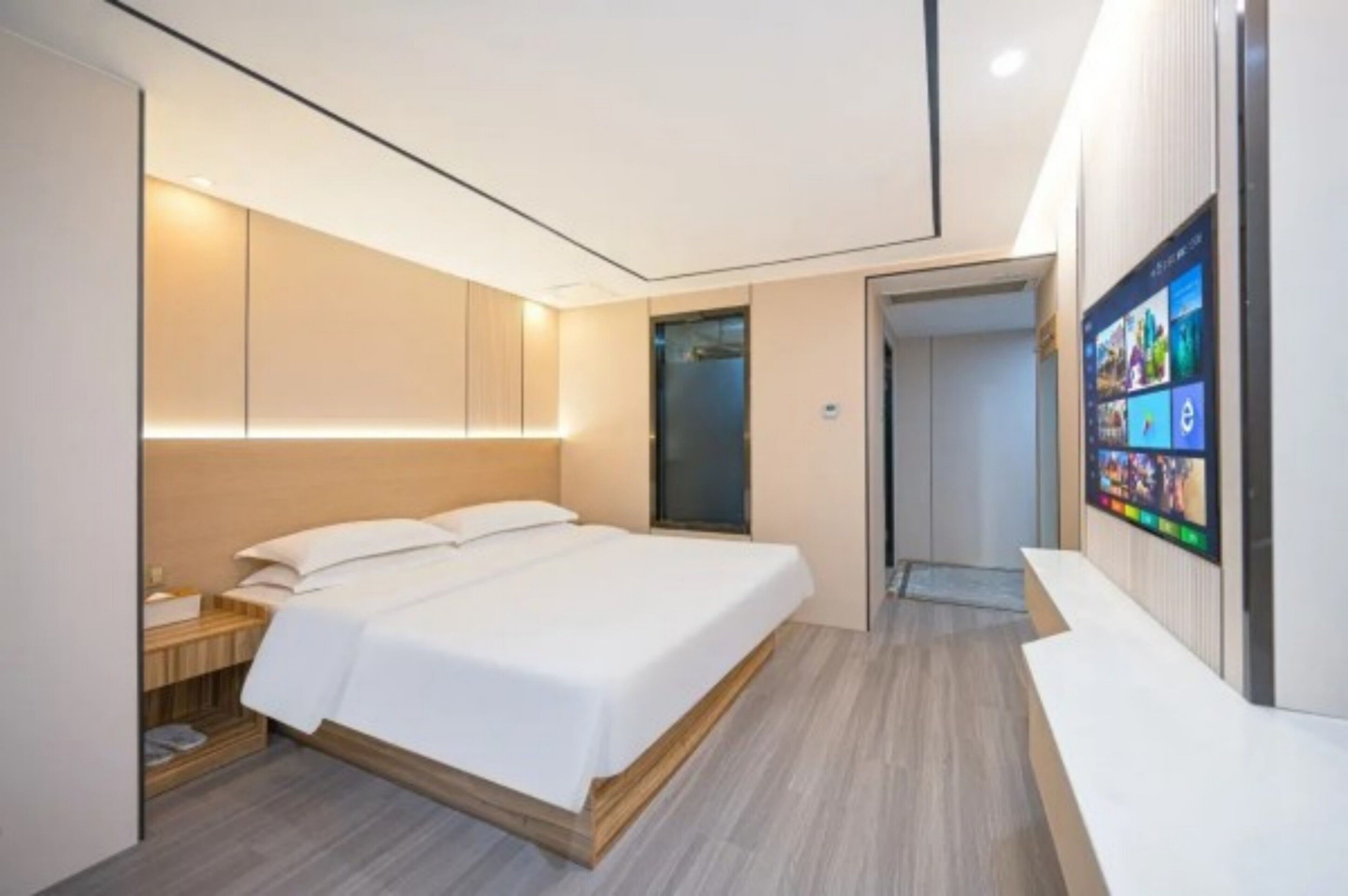 A modern hotel room with a large bed, a bedside table, a TV mounted on the wall, and a wooden floor.