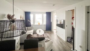 Ein modernes Wohnzimmer mit einer Couch, einem Couchtisch und einem Esstisch. Ein großes Fenster mit Vorhängen, ein Fernseher und eine Tür zu einem anderen Raum sind ebenfalls vorhanden.