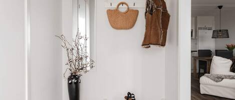 A hallway with a woven basket, a brown jacket, a black vase with branches, and a pair of black sandals on a metal bench.