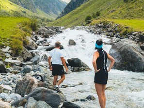 Deux personnes se tiennent sur des rochers, au bord d’une rivière où l’eau coule, avec des montagnes en arrière-plan.