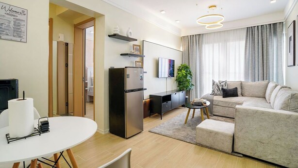 A modern living room with a sofa, a dining table, a kitchen area with a refrigerator, and a TV. - Modern 1BR Near JKIA & SGR | Pool, Gym & Free Parking (Nairobi)