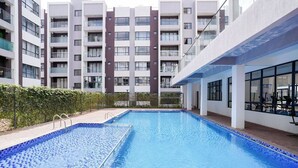 A swimming pool with a tiled border, surrounded by a hedge, and adjacent to a multi-story building with balconies. - Modern 1BR Near JKIA & SGR | Pool, Gym & Free Parking (Nairobi)