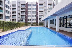 A swimming pool with a tiled border, surrounded by a hedge, and adjacent to a multi-story building with balconies. - Modern 1BR Near JKIA & SGR | Pool, Gym & Free Parking (Nairobi)