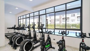 A gym with stationary bikes, treadmills, and elliptical machines, large windows, and a view of a building and greenery. - Modern 1BR Near JKIA & SGR | Pool, Gym & Free Parking (Nairobi)