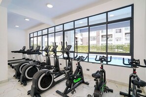 A gym with stationary bikes, treadmills, and elliptical machines, large windows, and a view of a building and greenery. - Modern 1BR Near JKIA & SGR | Pool, Gym & Free Parking (Nairobi)