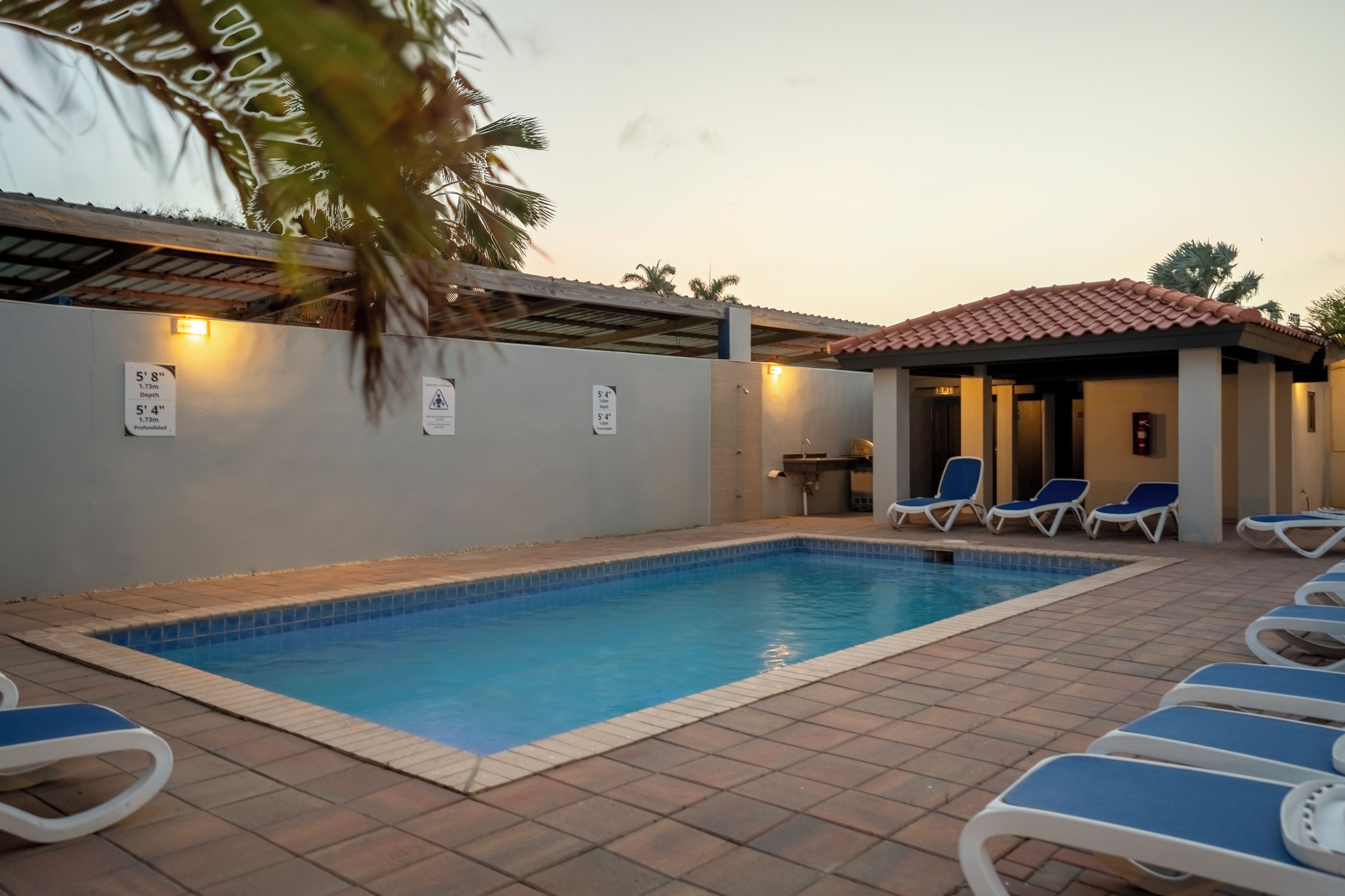 A swimming pool with blue tiles, surrounded by lounge chairs and a covered area with a tiled roof.