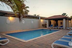 A swimming pool with blue tiles, surrounded by lounge chairs and a covered area with a tiled roof.