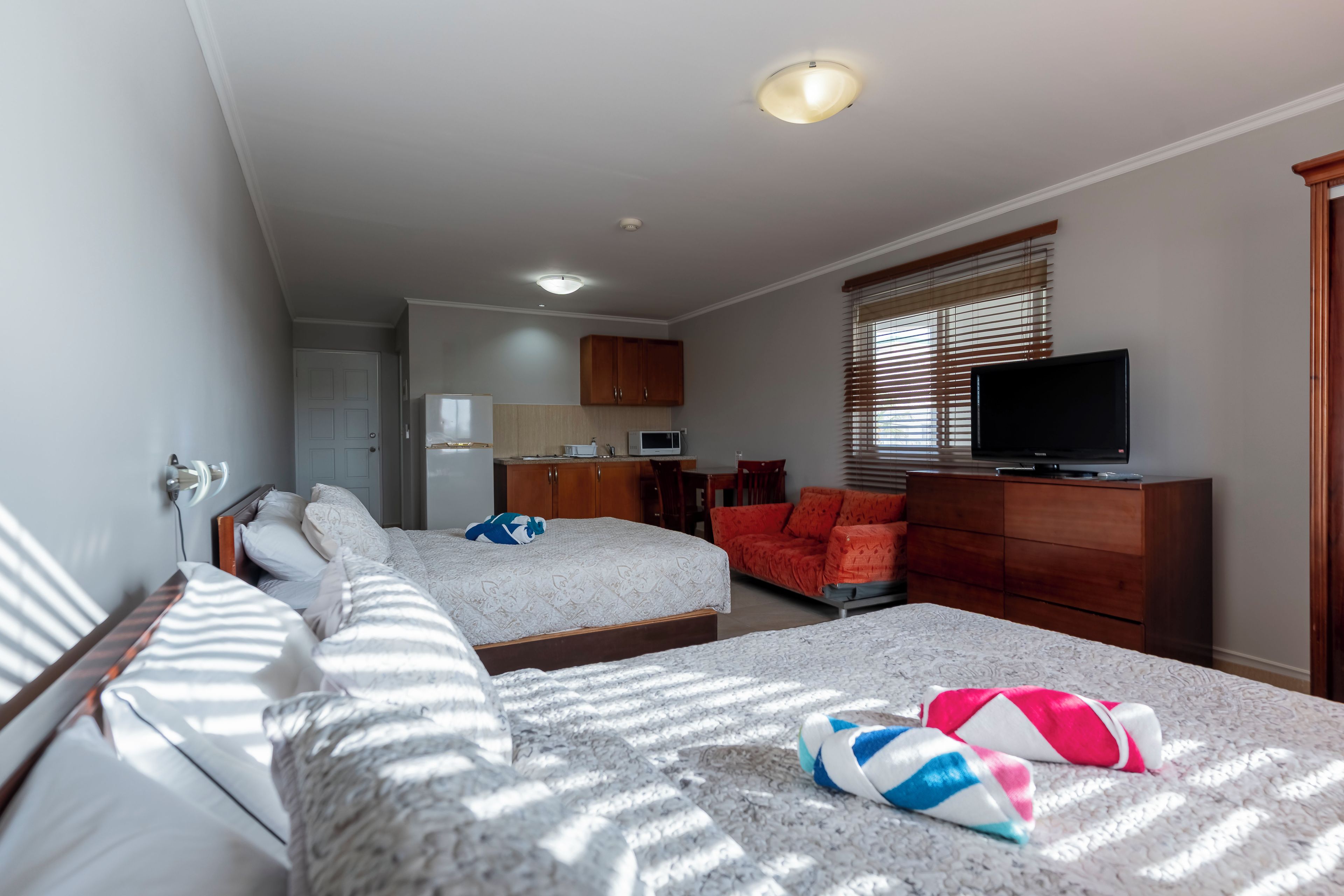 A hotel room with two beds, a television, a desk, and a kitchenette.