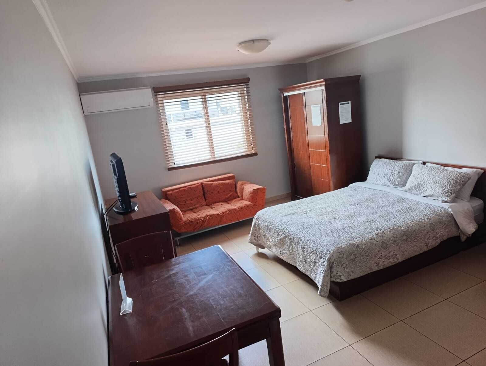 A bedroom with a bed, a desk, a chair, a wardrobe, and a window with blinds.