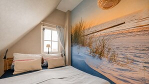 Une chambre à coucher avec un lit, des oreillers, une table de chevet, une lampe, une fenêtre avec des rideaux et une fresque murale représentant une plage.
