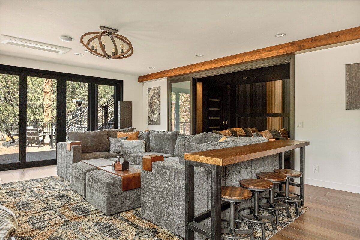 A modern living space with a large gray sectional sofa, a wooden bar with stools, and a view through large sliding glass doors.