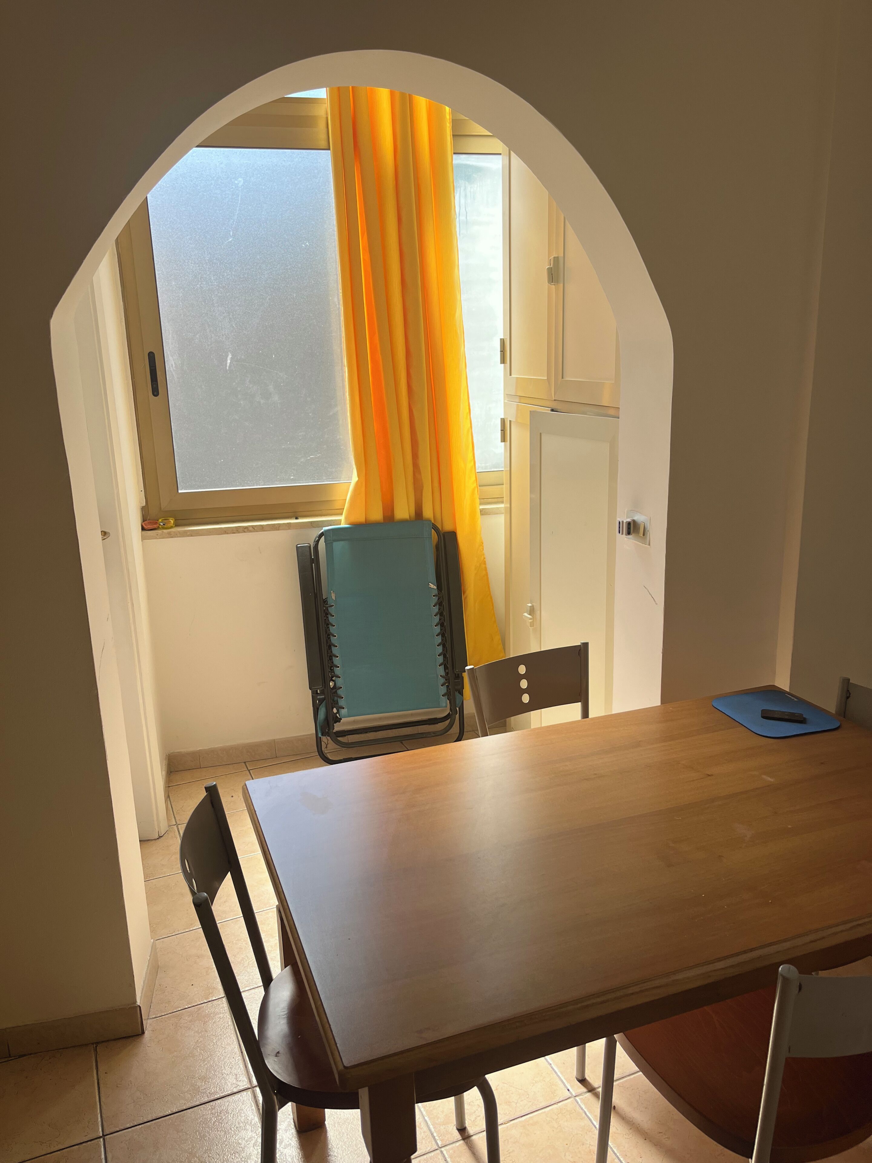 Ein Zimmer mit einem Holztisch, zwei Stühlen, einem Fenster mit gelben Vorhängen und einer Tür.