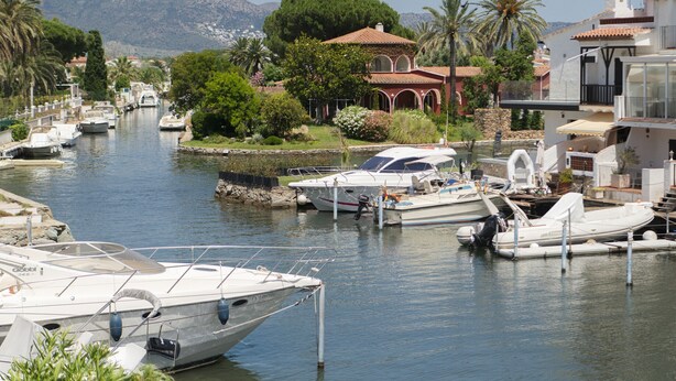 Un puerto deportivo con barcos amarrados, un edificio de techo rojo y montañas al fondo.