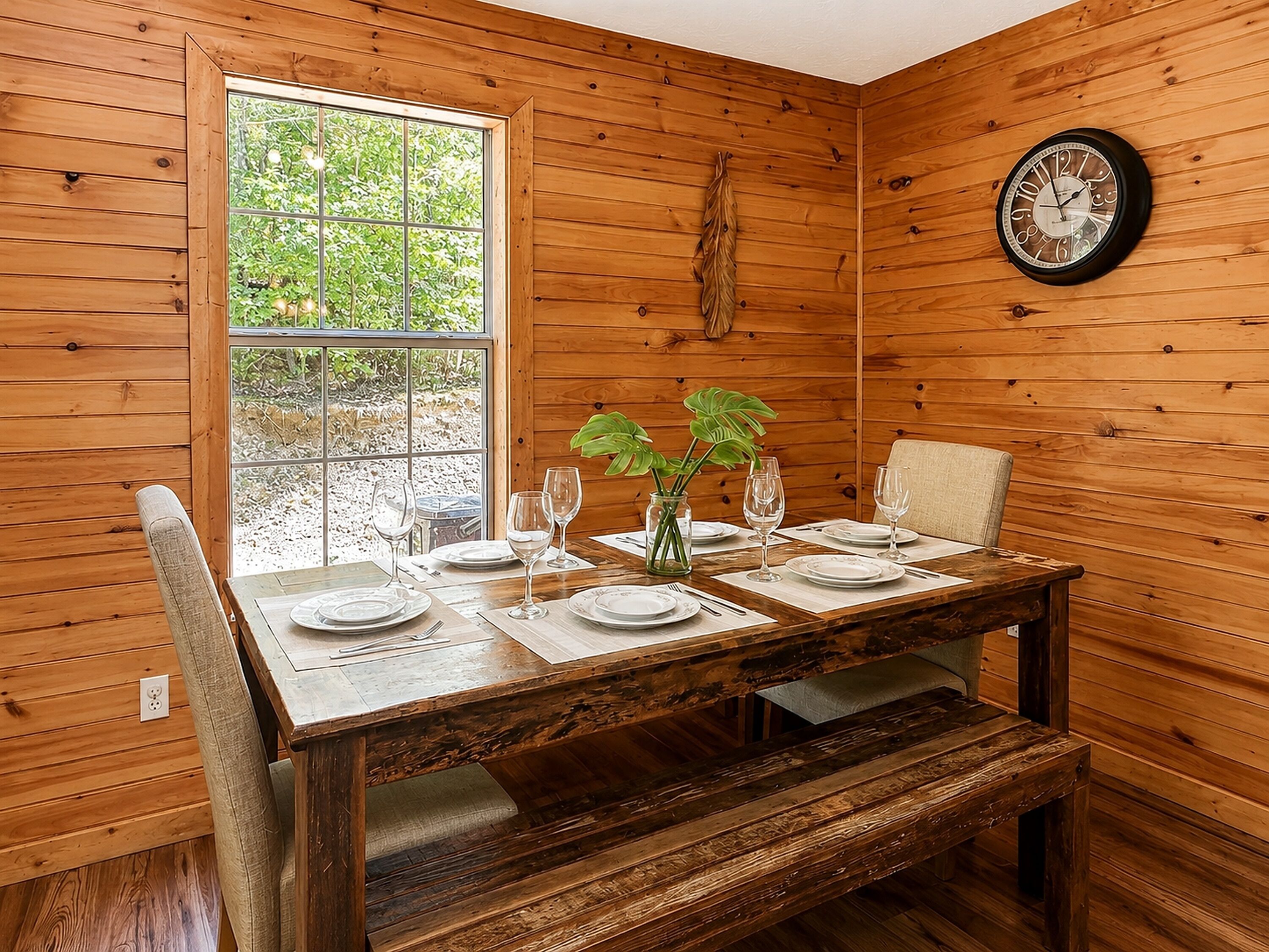 A wooden dining room with a table set for two, a clock on the wall, and a window with a view of greenery.