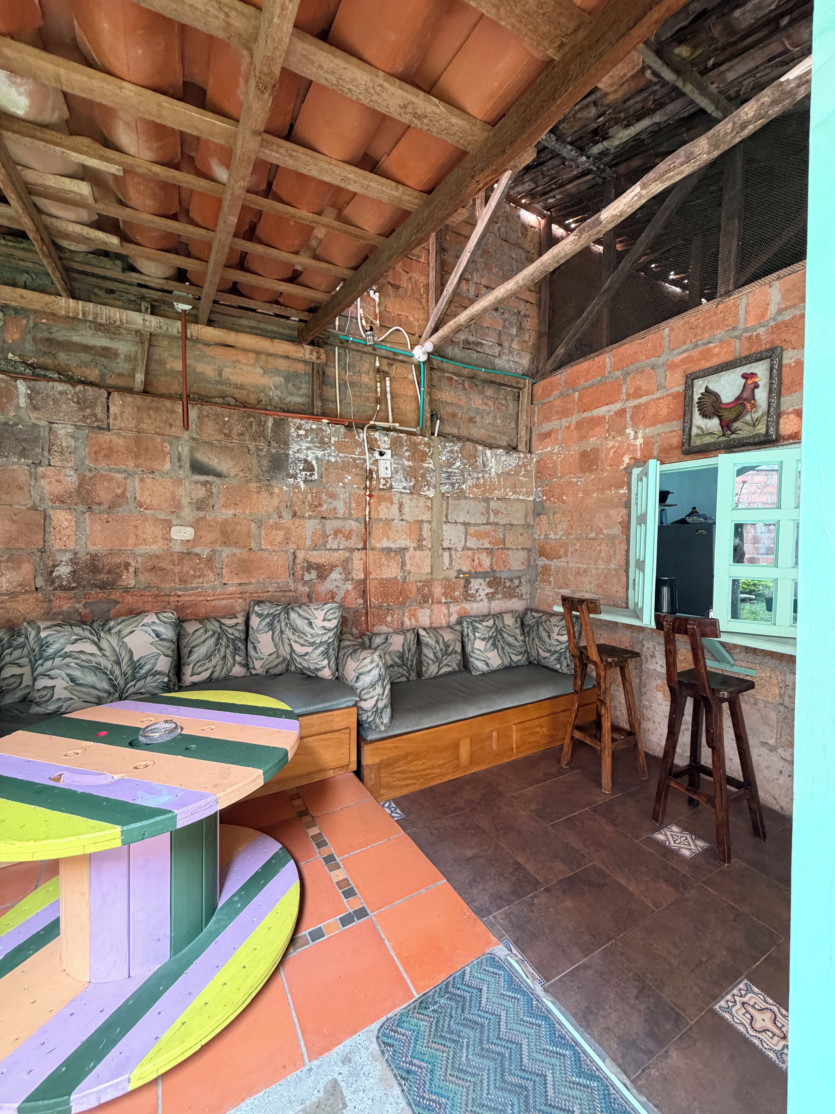 A rustic interior with a brick wall, wooden ceiling beams, a cushioned bench, a colorful table, and a window with a rooster decoration.