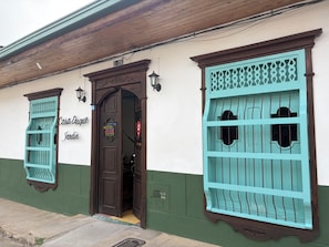 Un edificio con fachada verde y blanca, una puerta de madera y un letrero encima, y dos ventanas con rejas decorativas de metal.