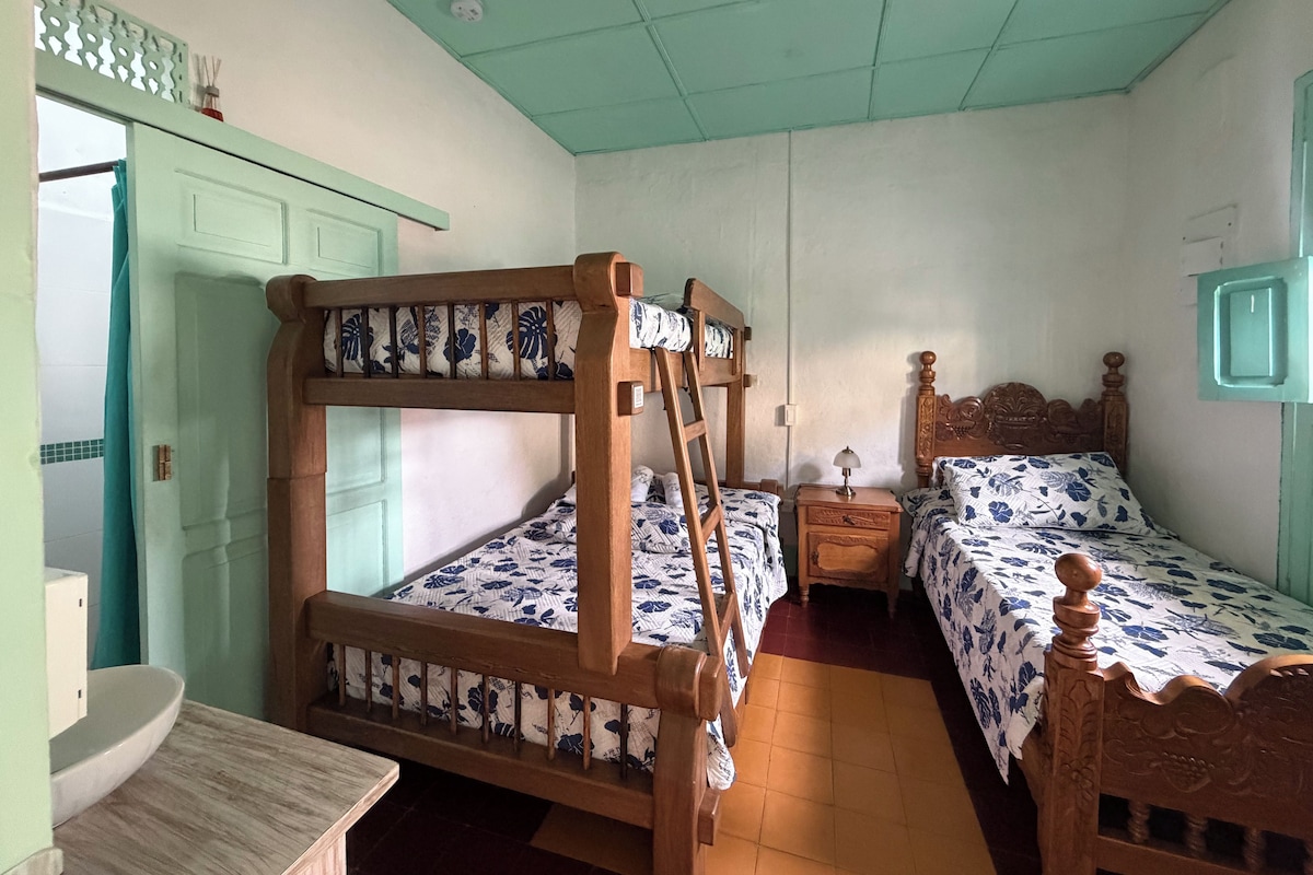 Una habitación con literas, una mesita de noche con lámpara, un armario pequeño y una ventana con cortinas.