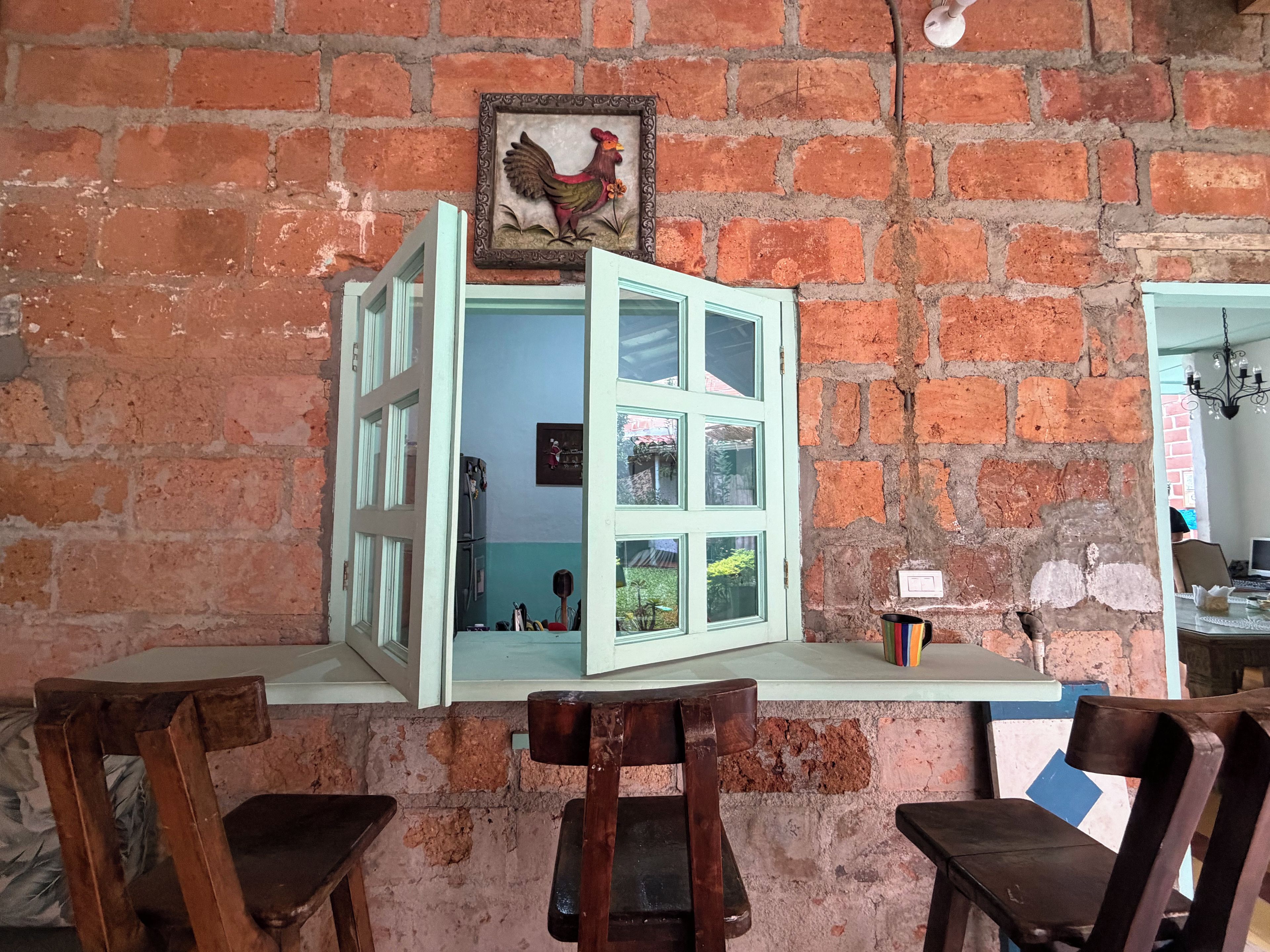 A rustic brick wall with a framed rooster decoration, a partially open green-framed window, and a small table with a cup.