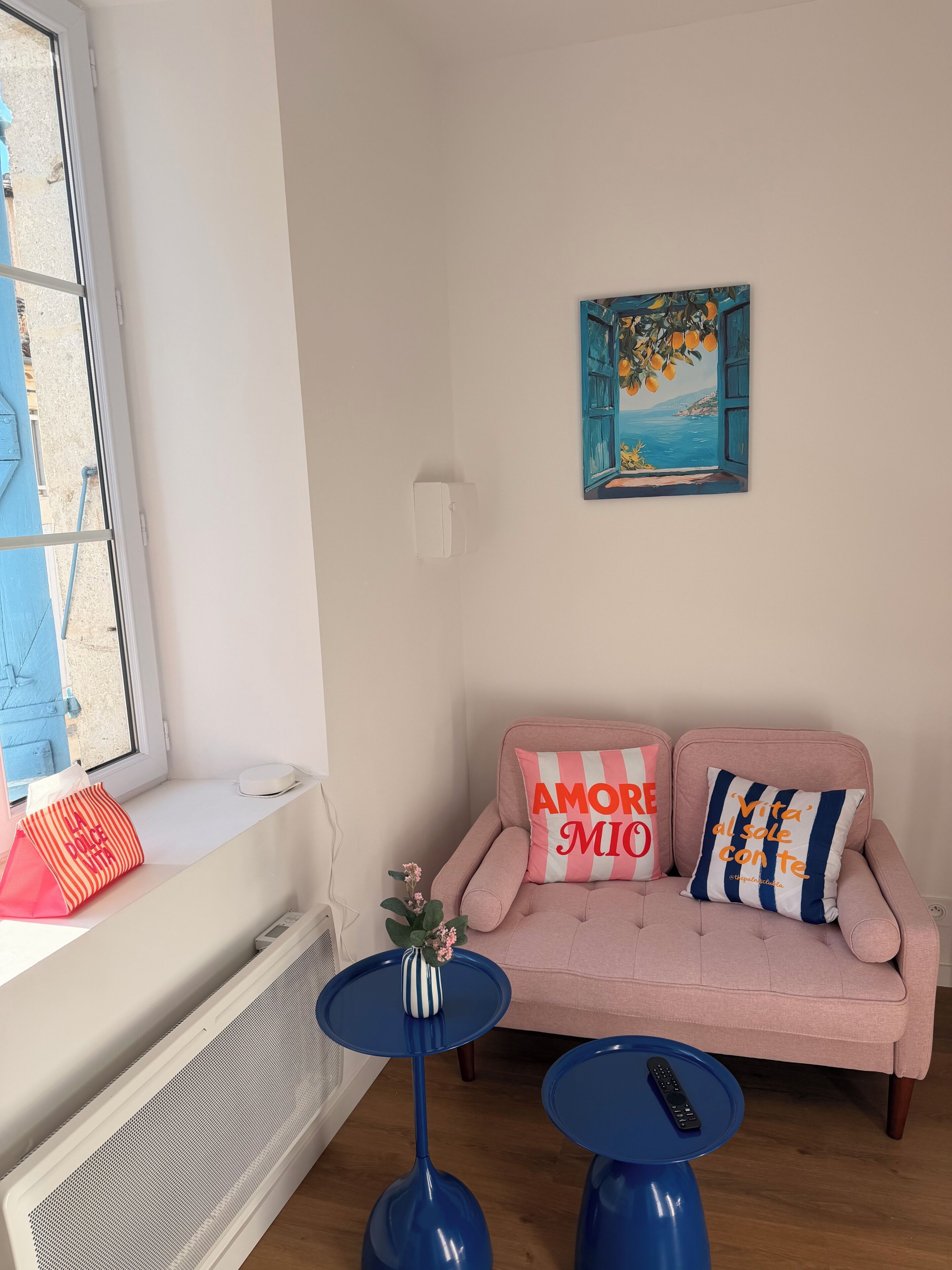 Un salon avec un canapé rose, deux coussins, une petite table, une table bleue, une fenêtre donnant sur un beau paysage et un tableau accroché au mur.