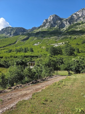 A mountainous landscape with a dirt path, greenery, and a building partially obscured by trees.