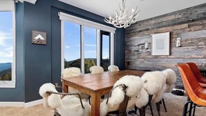 A dining area with a wooden table and white furry chairs, a chandelier, and a view through large windows. - Wintergreen Ridgeline Retreat (Wintergreen Resort)