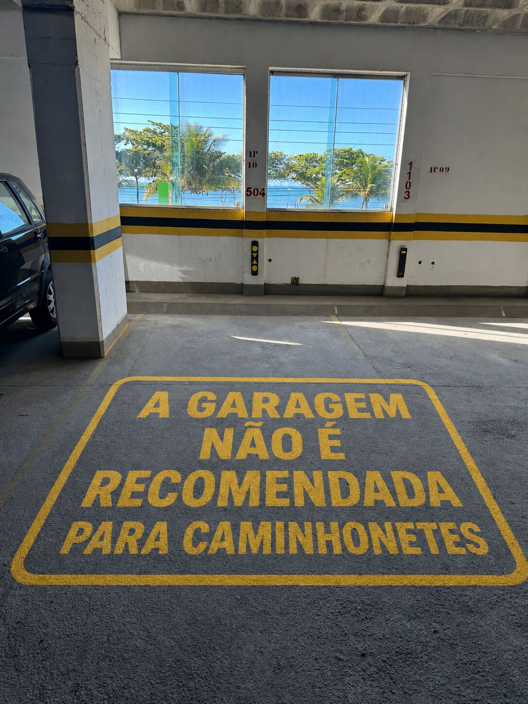 Un garaje de estacionamiento con franjas amarillas y negras, un automóvil estacionado en su interior y un letrero en portugués que advierte contra el estacionamiento de motocicletas.