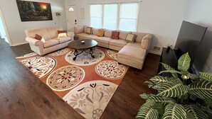 Una sala de estar con un sofá seccional, una mesa de centro, un tapete estampado, una planta grande y un televisor.