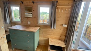 A wooden cabin interior with a green dresser, two windows with curtains, a small table, and a sliding glass door.