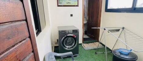 A laundry area with a washing machine, a drying rack, a chair, and a trash bin.