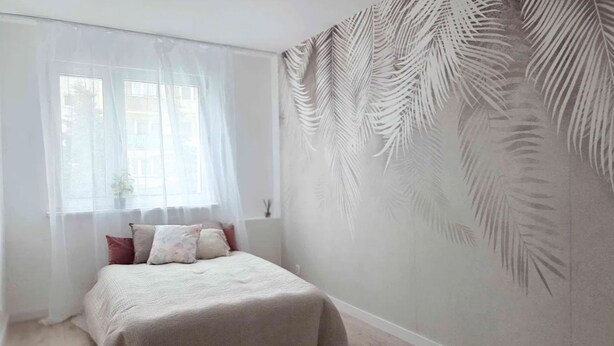A bedroom with a bed, a window with sheer curtains, and a wall with a palm leaf pattern.