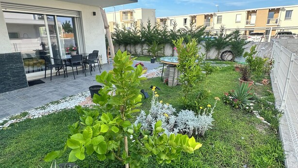 Een modern terras buiten, met glazen deuropening, een zithoek en een verzorgde tuin met diverse planten en een kleine waterpartij.
