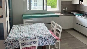 Een keuken met een eettafel, stoelen, een raam met jaloezieën en een koelkast.