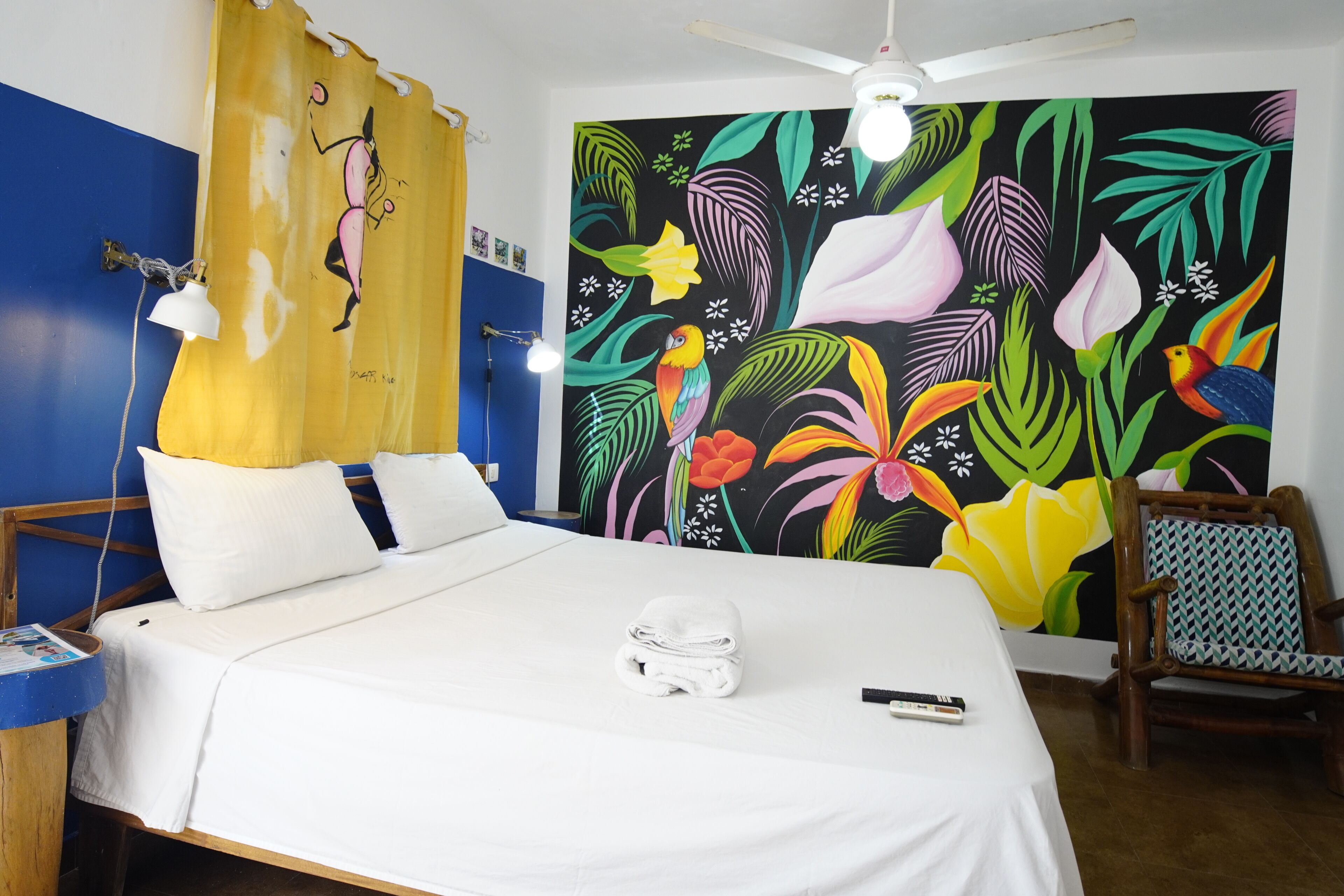 Una habitación de hotel con cama, una silla, un mural en la pared y un ventilador de techo.
