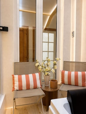 Ein Zimmer mit zwei Sitzbereichen, jeweils mit Kissen und einem kleinen Tisch. Im Hintergrund ist ein Fenster mit Milchglas und eine Holztür zu sehen.
