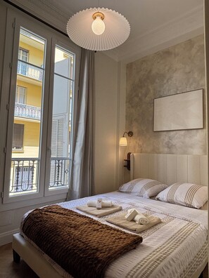 Ein Hotelzimmer mit einem großen Bett, einer Nachttischlampe, Blick auf einen Balkon und einem Wandbild.