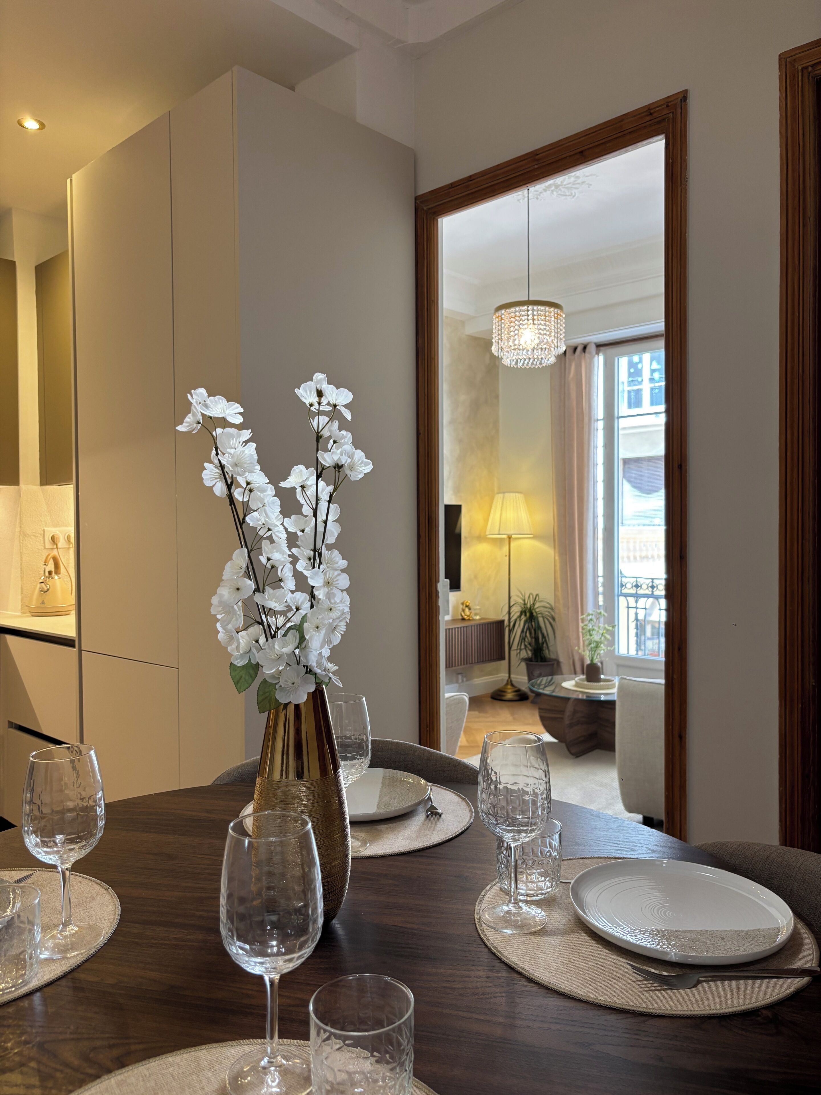 A dining area with a table set for four, a vase with white flowers, and a view into a living room.