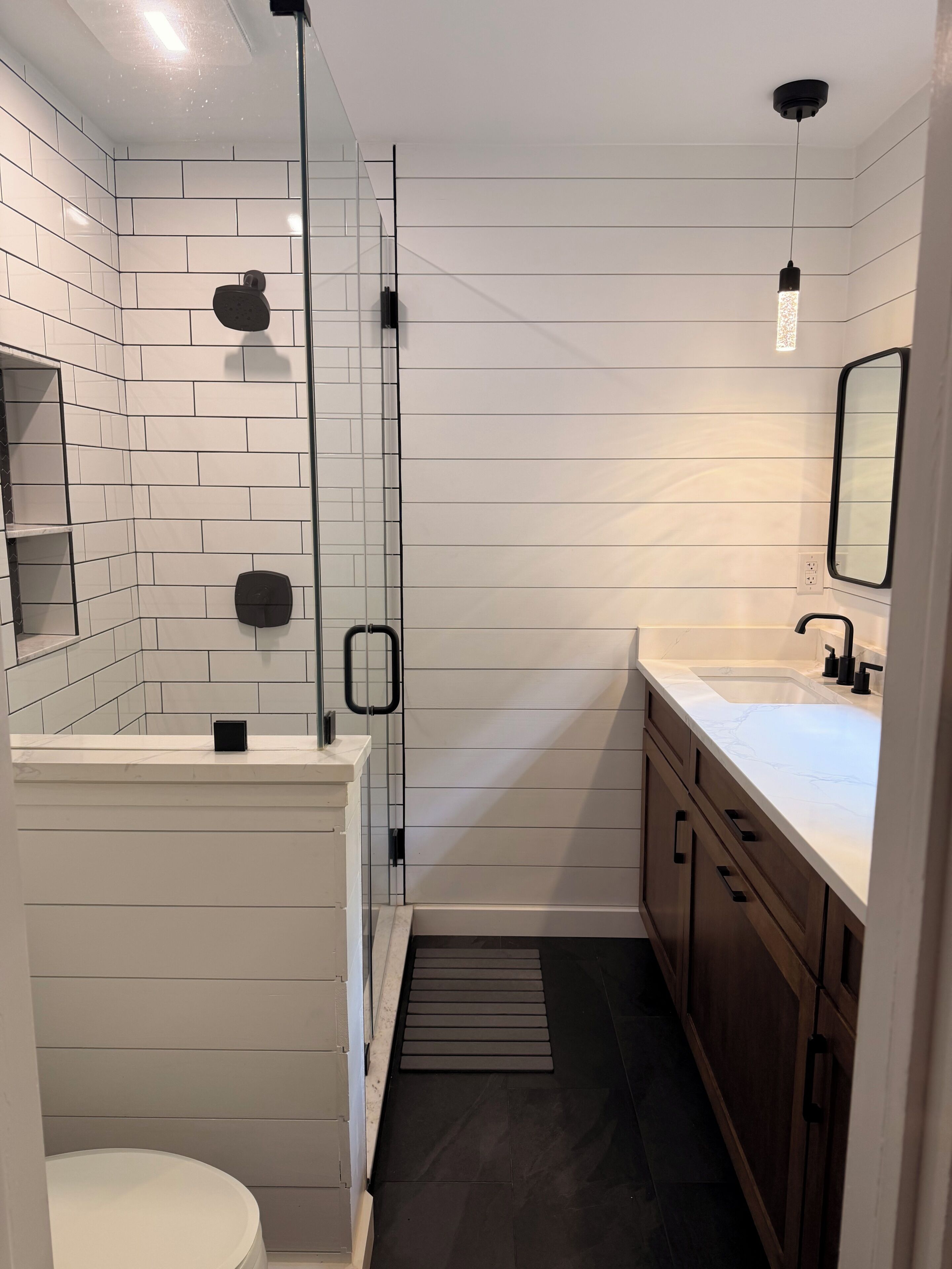 A modern bathroom with white subway tiles, a glass shower enclosure, a white toilet, wooden cabinets, and a marble countertop.