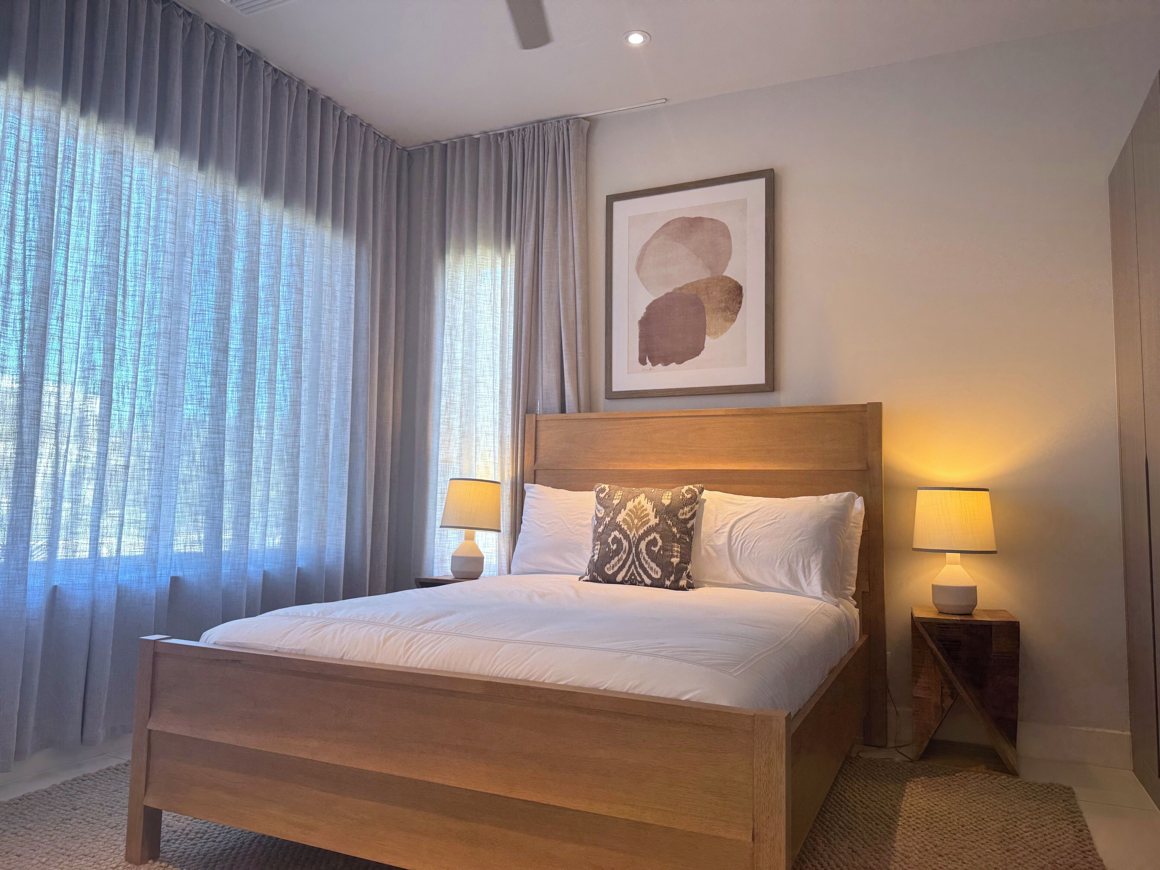 A bedroom with a wooden bed, two bedside lamps, a large window with sheer curtains, and a framed abstract painting on the wall.