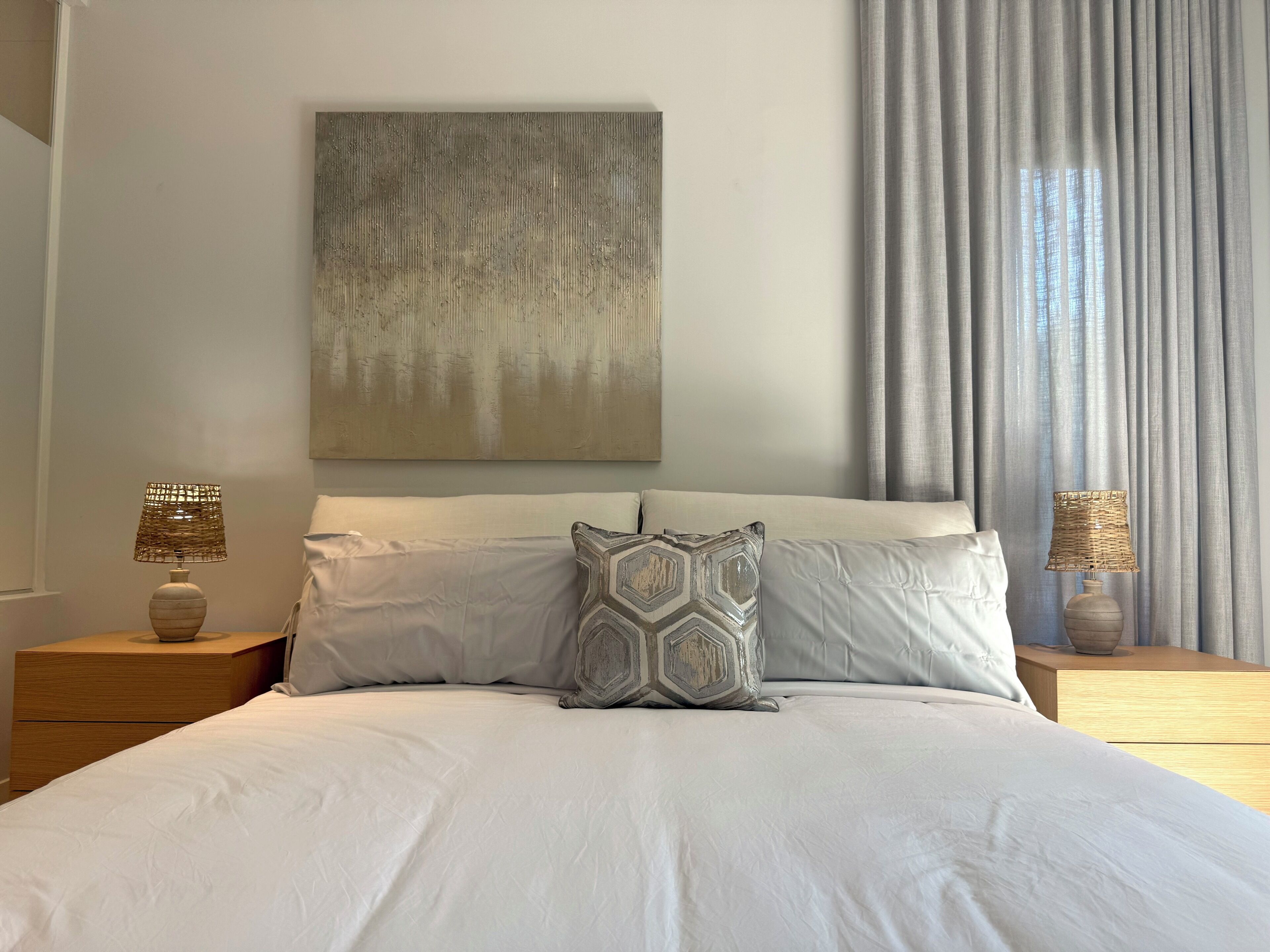 A neatly made bed with white linens, two bedside tables with lamps, a geometric-patterned pillow, and a large abstract painting on the wall.